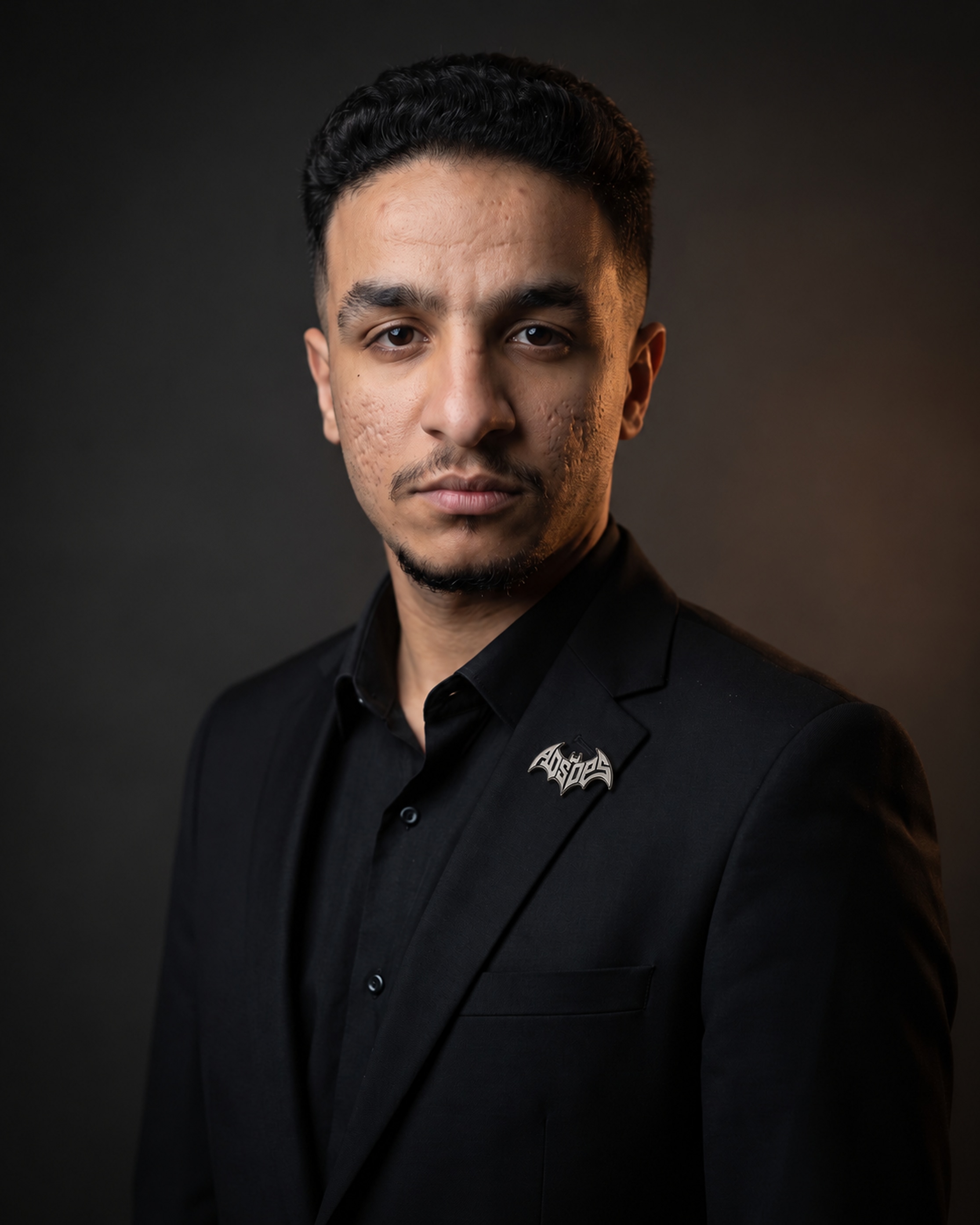 Aser Agina in a black suit and black shirt against a charcoal background, looking directly at the camera.