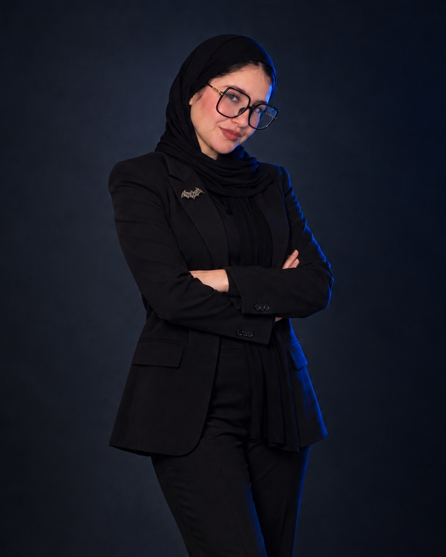 Rawan El-Mallah in a black suit and black hijab, arms folded, against a deep blue-grey background.