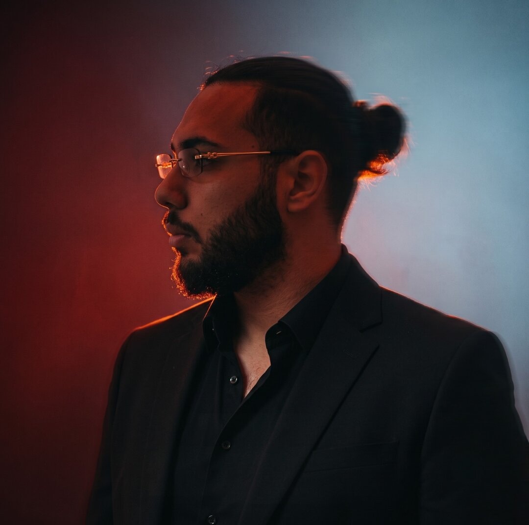 Yousef Abdo in profile, wearing glasses and a black suit, with red and blue lighting.